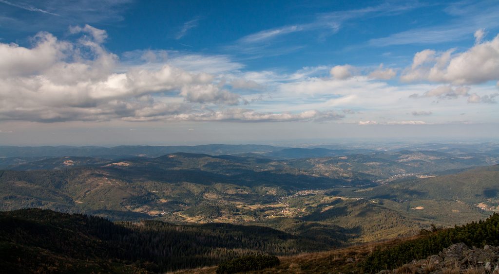 Panorama beskidzkich wzgórz i dolin z rozproszoną zabudową, nad nimi błękitne niebo z chmurami.
